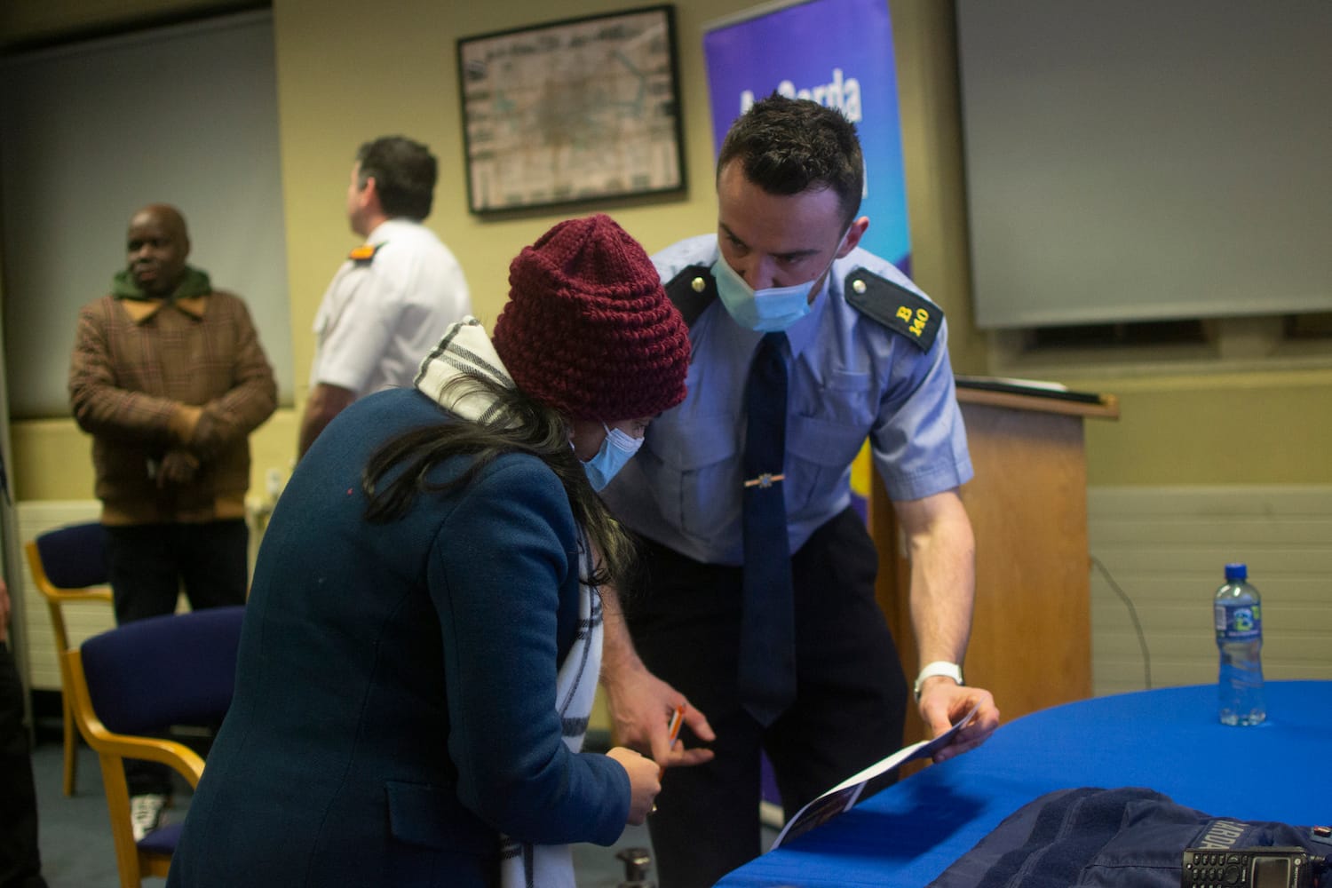 A garda in a face mask is talking to a woman in a woolly hat. They are inside a conference room. Two other people are stood in the background.