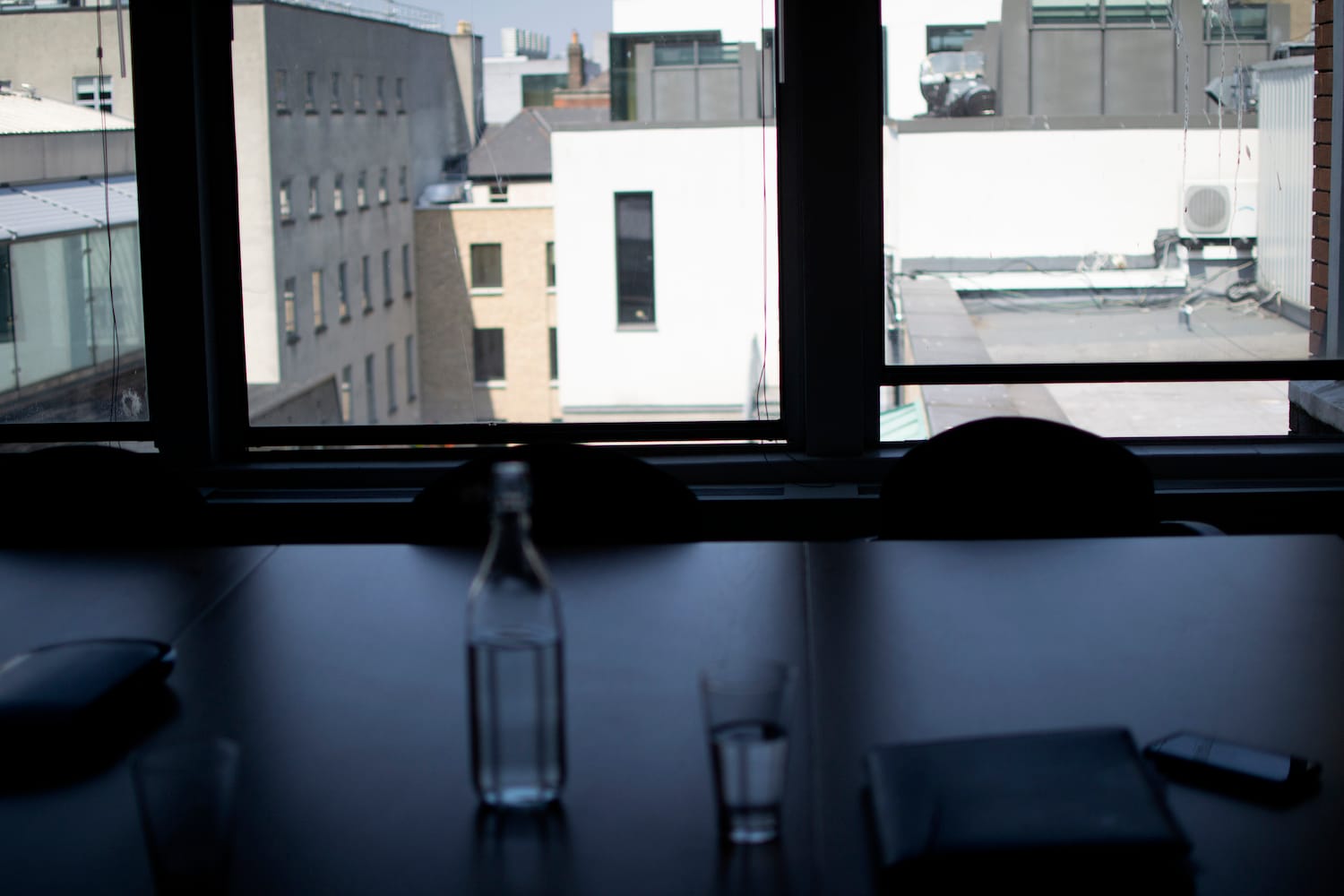 A glass water bottle on a big table. In the background is a big window, looking out onto blocky buildings.