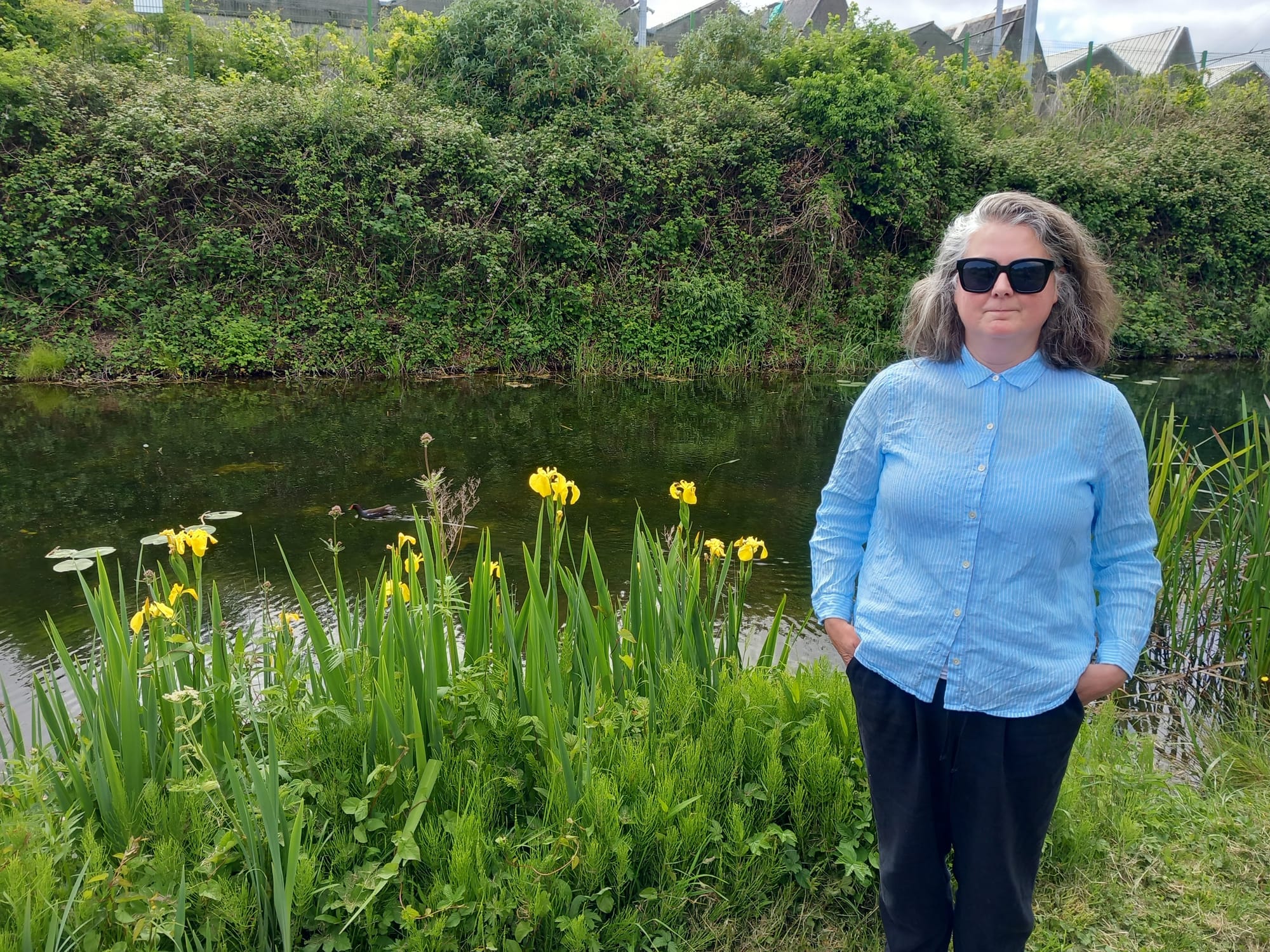 A woman in a blue jacket adn black sunglasses stands to the right of the image, with her hands in her pockets. She stands with her back to the canal, and a patch of yellow flowers.