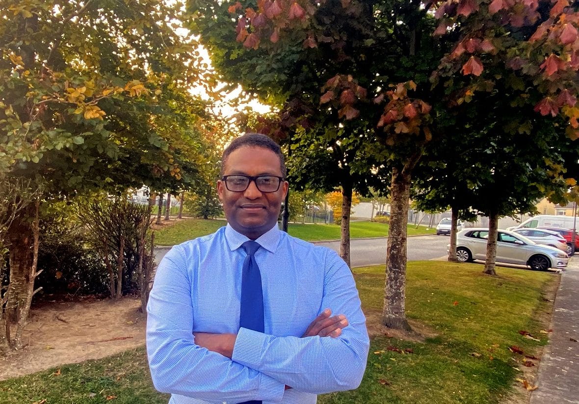 An image of a man in a blue shirt and tie, against a backdrop of trees. 