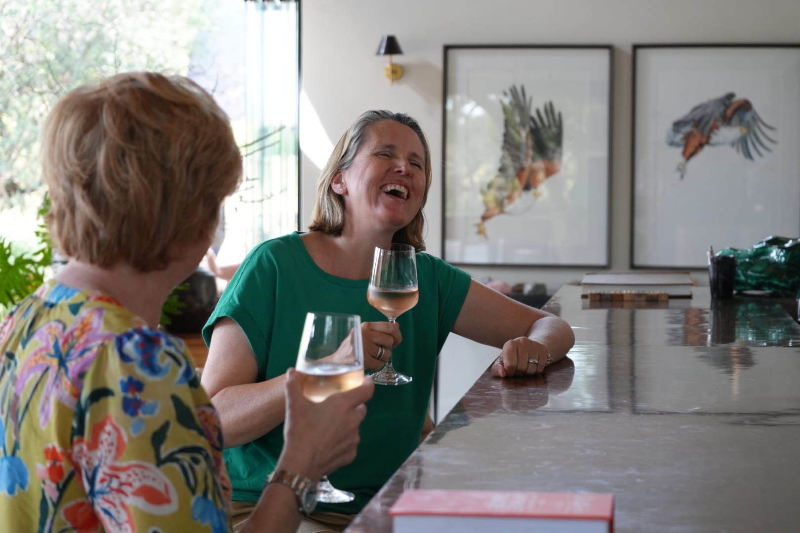 Vikki and Sian laughing at the Monwana bar with glasses of rosé, framed prints of African harrier-hawks on the wall behind.