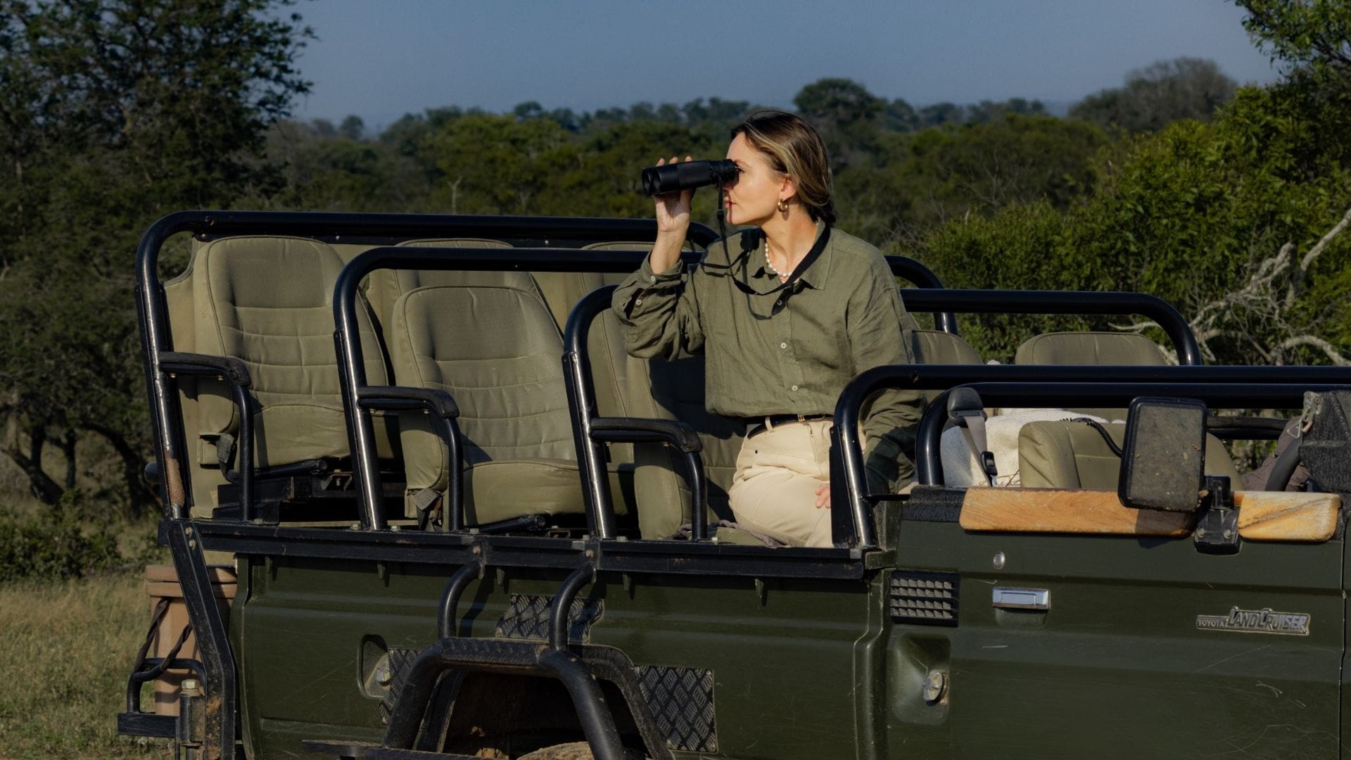 Solo traveller in a Land Cruiser, binoculars raised, empty seats around her, golden-hour bushveld