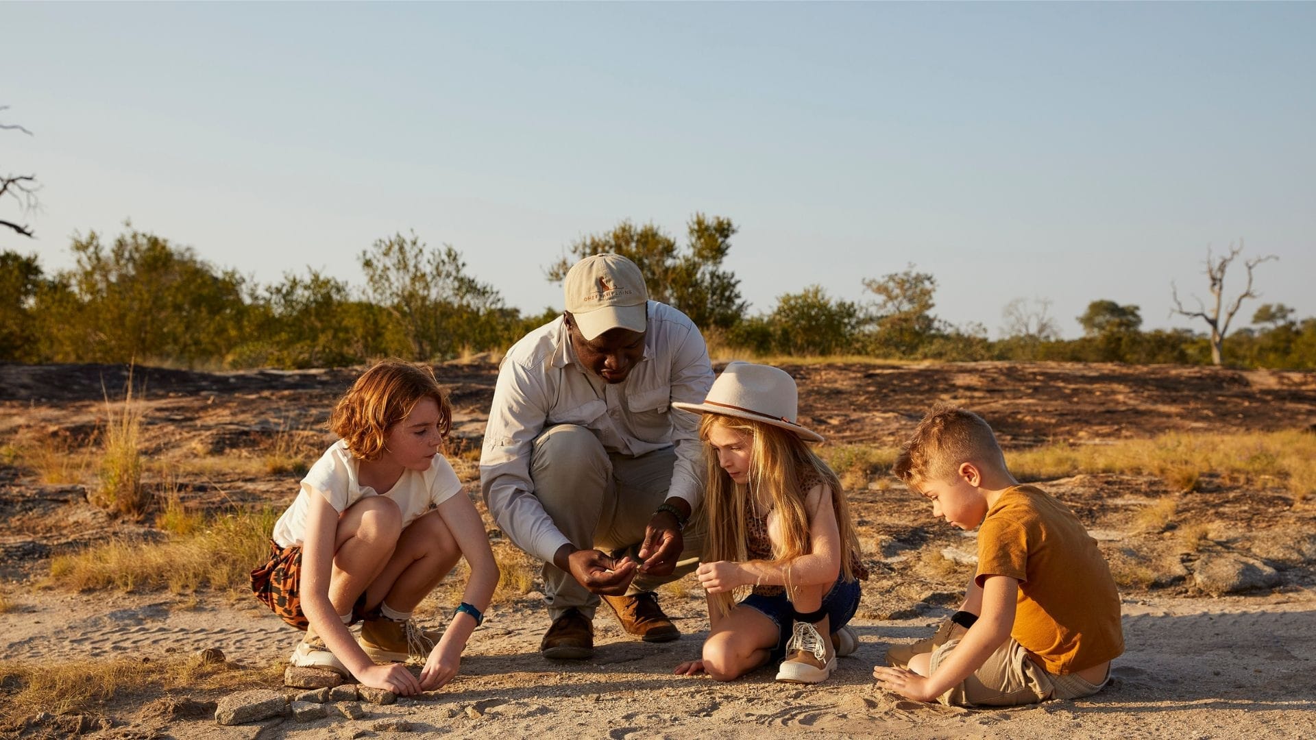 Guide crouched on the sand with three children examining tracks, late-afternoon bushveld light