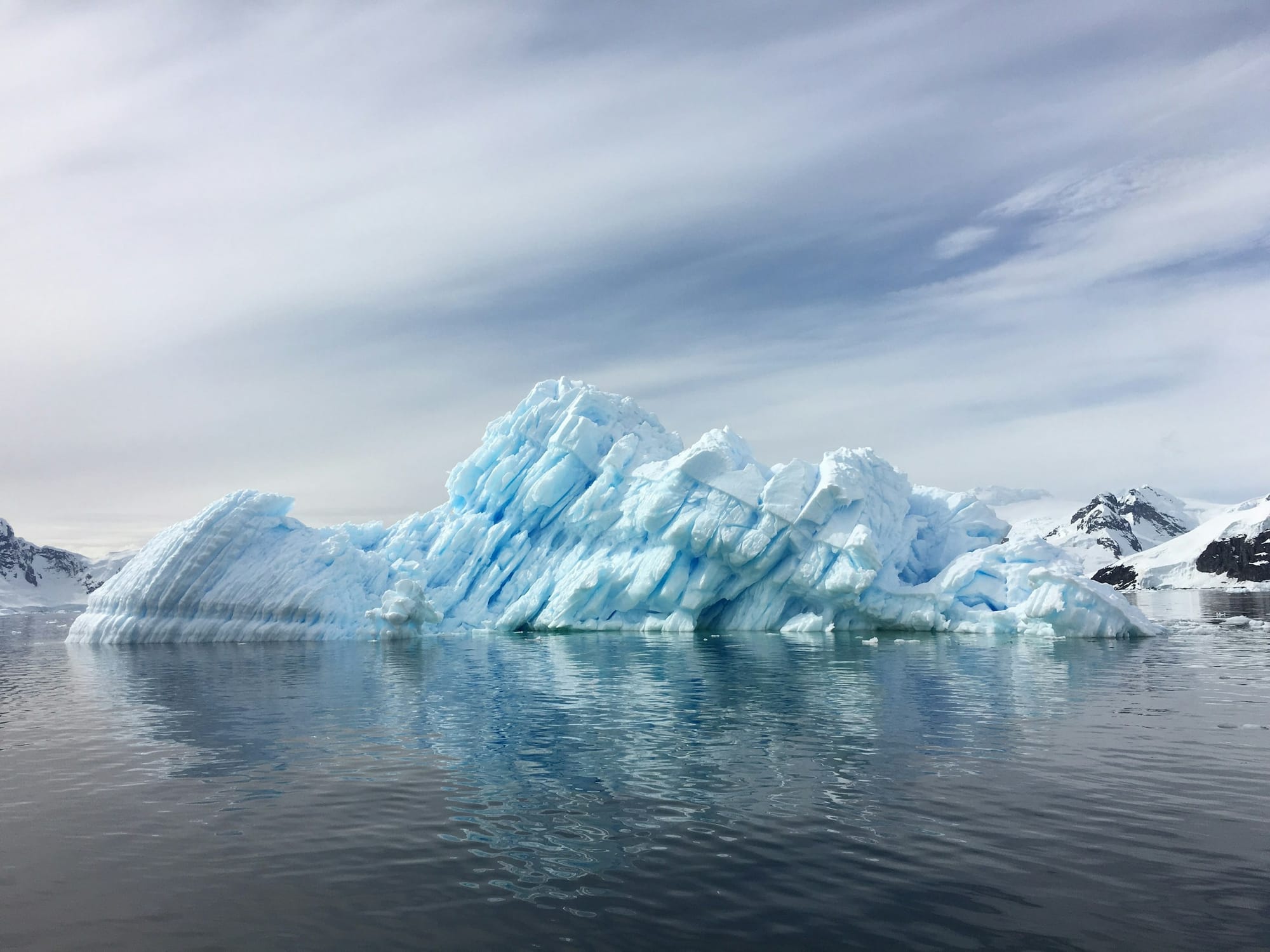 Sculptural pale-blue iceberg in calm Antarctic water, snow-capped mountains beyond, soft polar light overhead