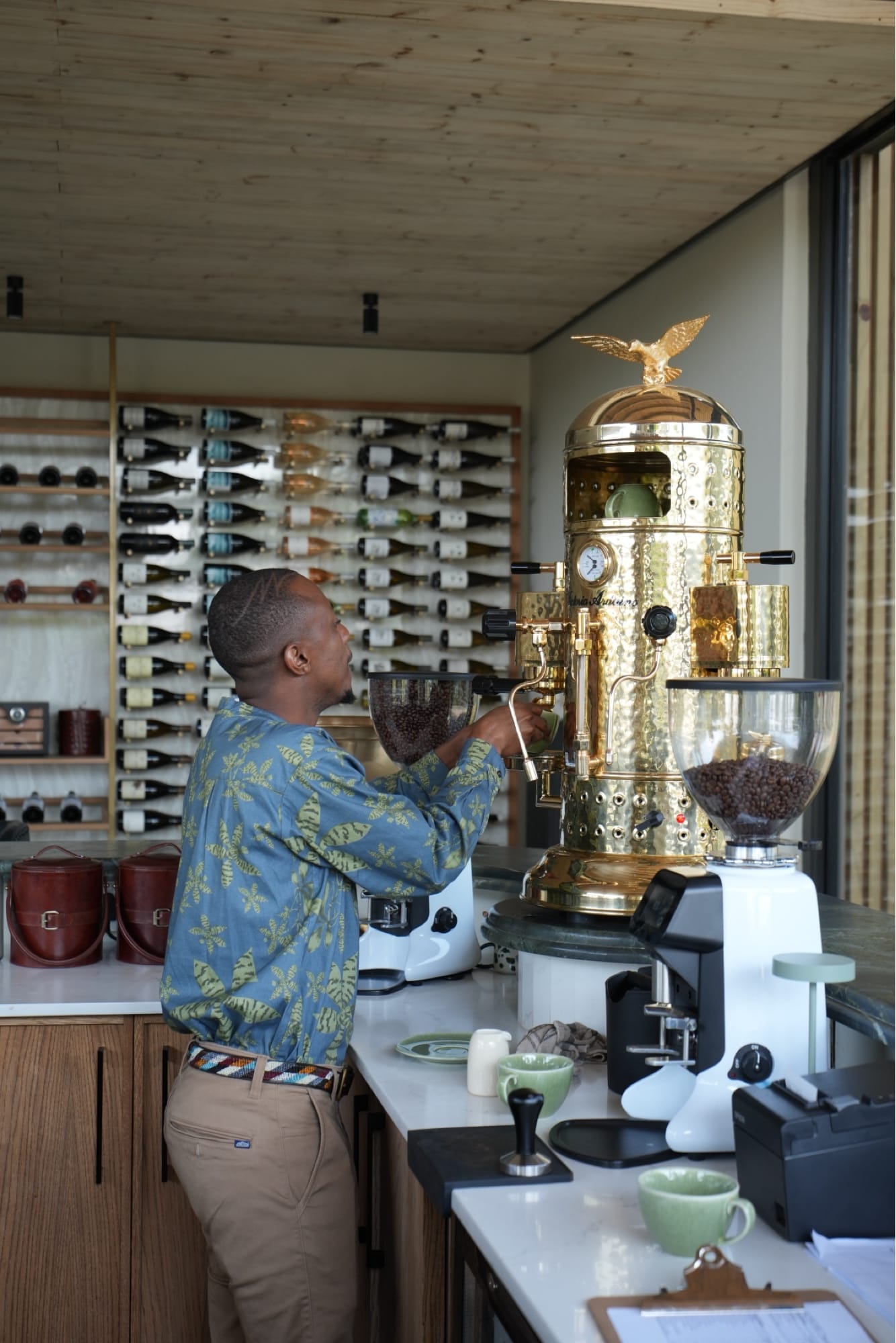 Barista at the gold espresso machine with the backlit wine wall behind, Lion Sands Ivory Lodge