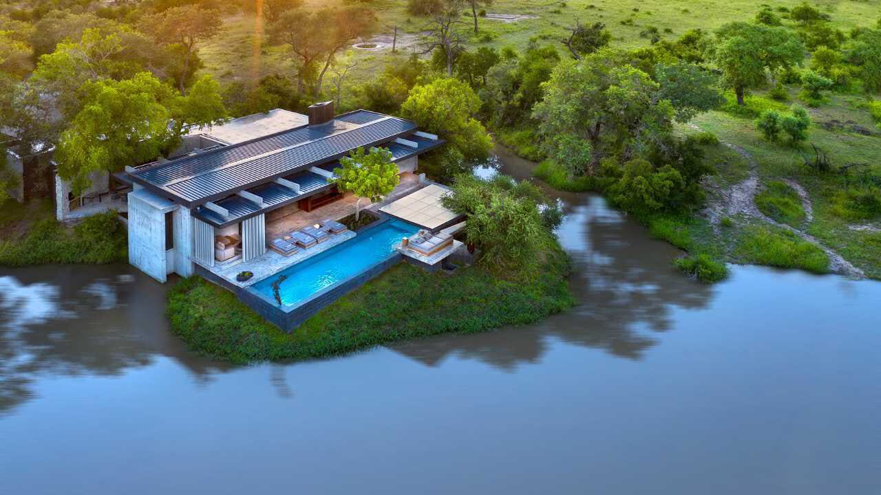 Aerial of a private safari villa with infinity pool overlooking a river, late-afternoon light, the pause point of a family itinerary