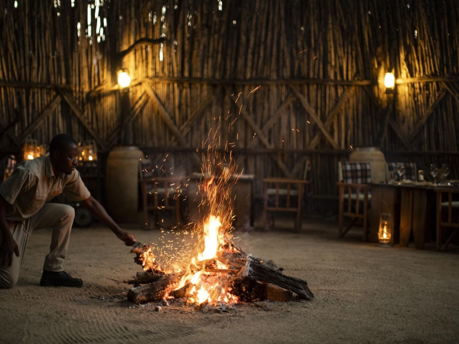 Chef tending a low fire in a reed boma, sparks rising, dining table set quietly behind, lanterns lighting the cane walls, the unfussy version of a bush dinner