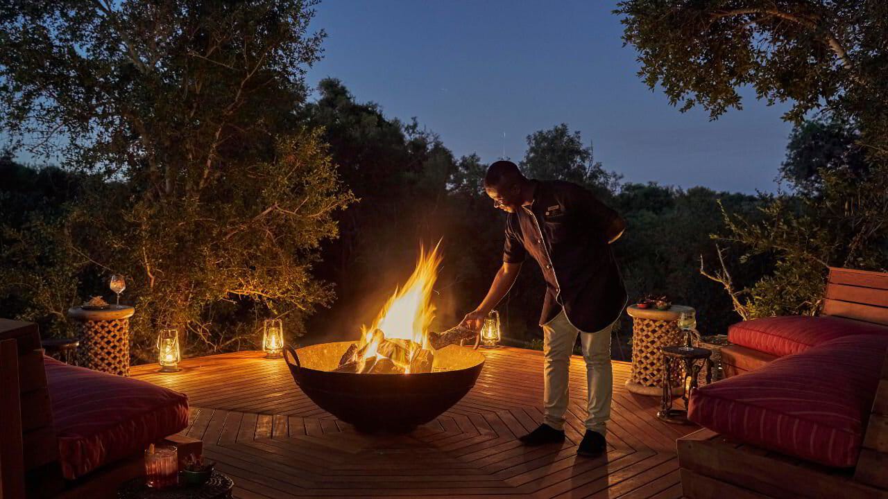 A safari lodge staff member tending a fire pit at dusk in the African bush
