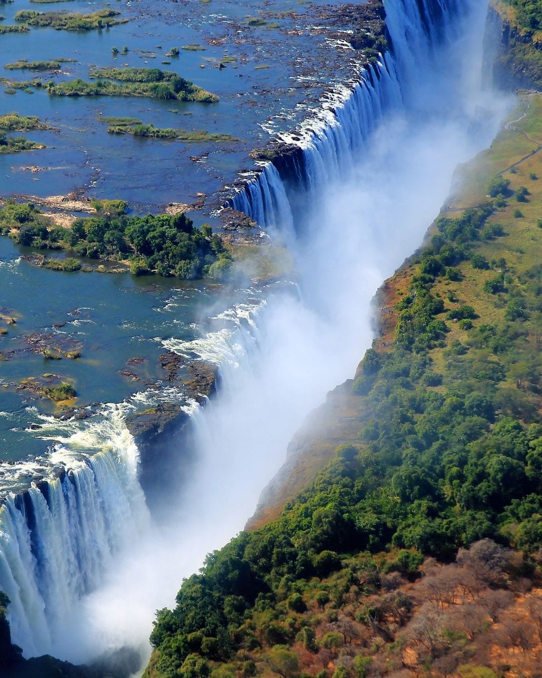 An aerial view of Victoria Falls in full flow, a long plume of mist rising as the Zambezi drops over the basalt edge with green riverine forest along the gorge