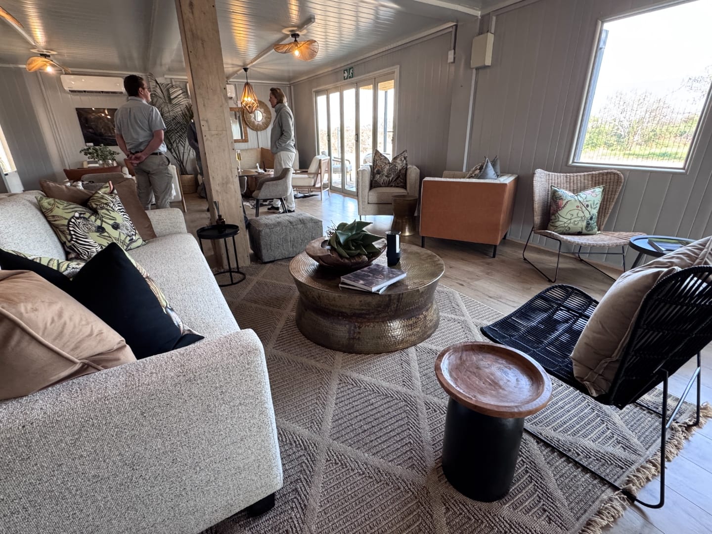 Cream upholstered seating, rattan armchairs and a hammered coffee table by a window onto Lowveld trees