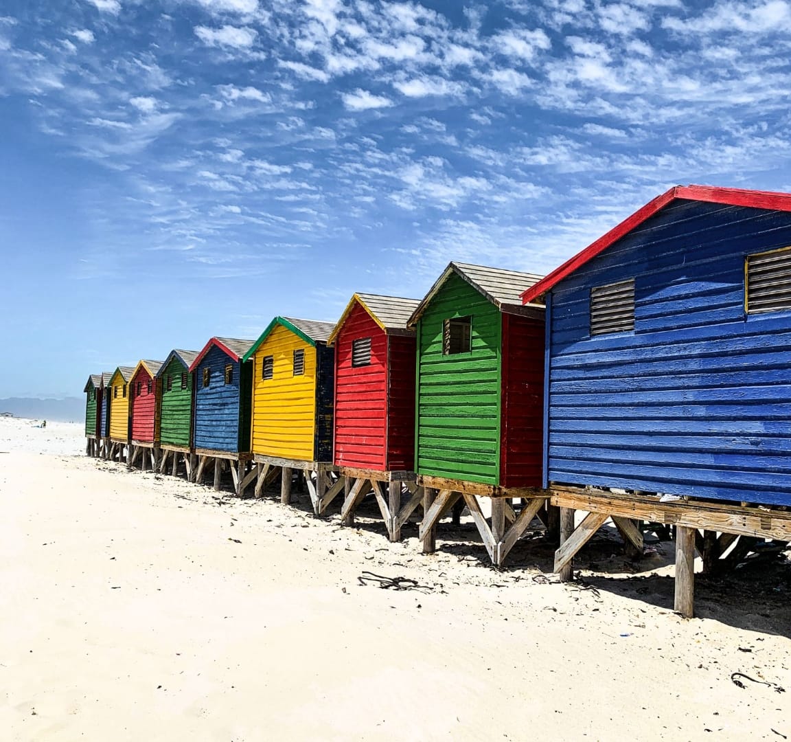 The painted Victorian bathing boxes at Muizenberg beach, Cape Town, lined up on white sand under a wide blue sky
