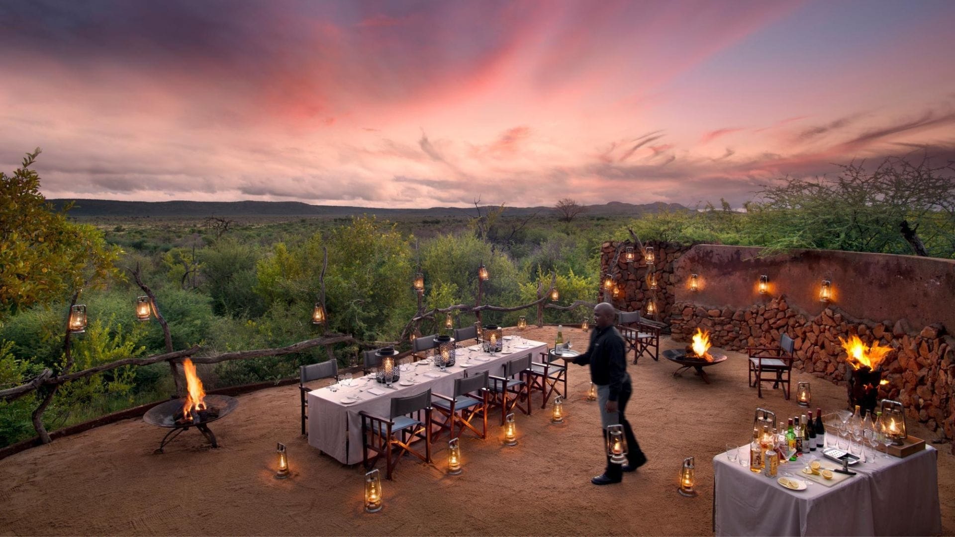 Family bush dinner under a pink sky in Madikwe, lanterns and a single staff member walking out with drinks