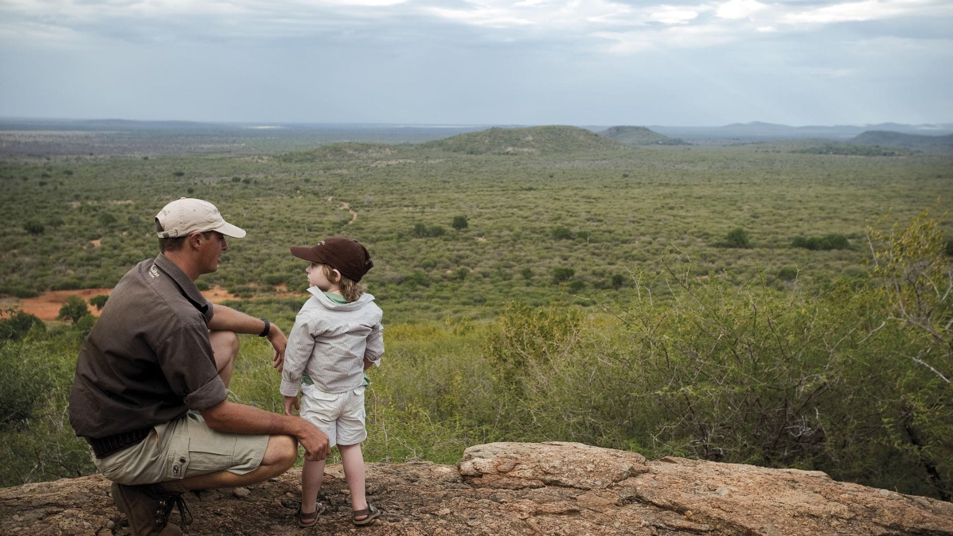 A ranger and a young child crouched on a rocky outcrop looking out over the South African bush