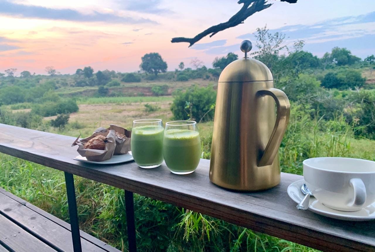 Two glasses of green juice, a brass plunger of coffee, a muffin and a cup on a deck above the Sabie River at sunrise