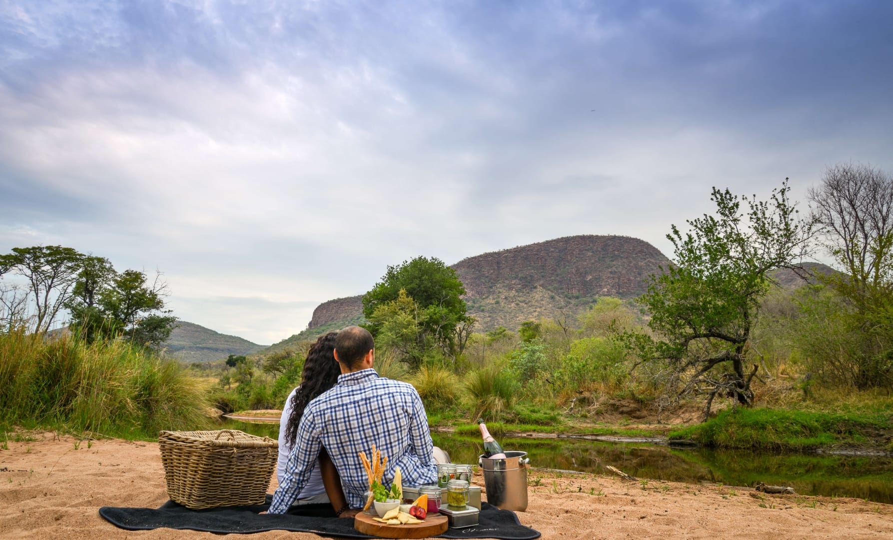 Couple picnicking on a blanket beside a river with acacia and baobab trees