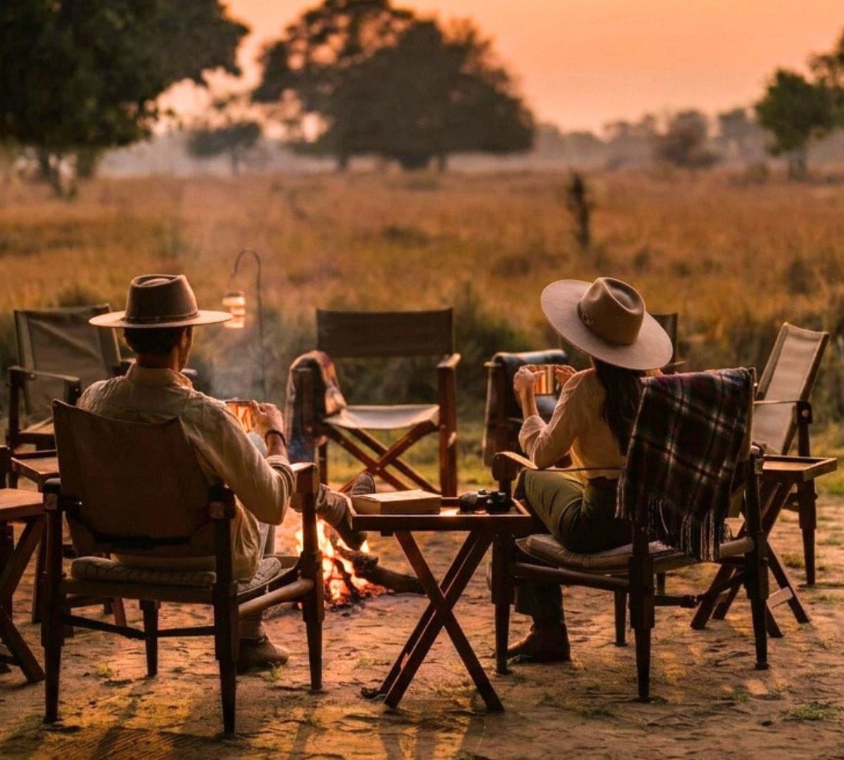 Two guests in safari hats sitting by a small fire at a properly small bush camp at golden hour, lantern lit, open floodplain stretching out behind