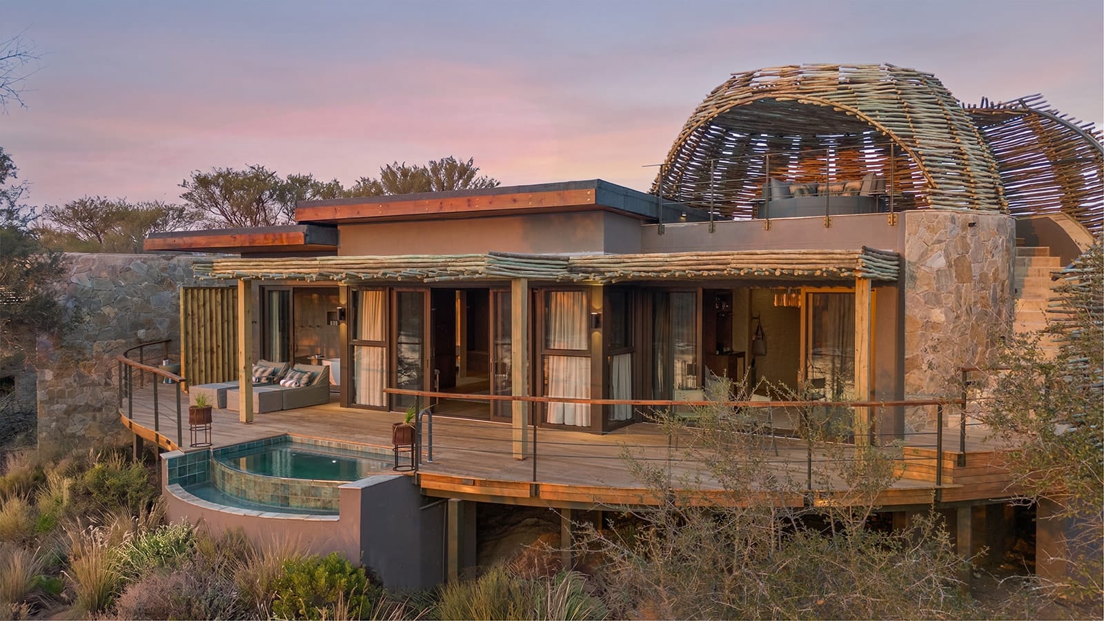 A curved timber deck with a private plunge pool outside a suite at Shisaka Camp, Sandringham, beneath a domed reed-and-timber rooftop lounge at dusk