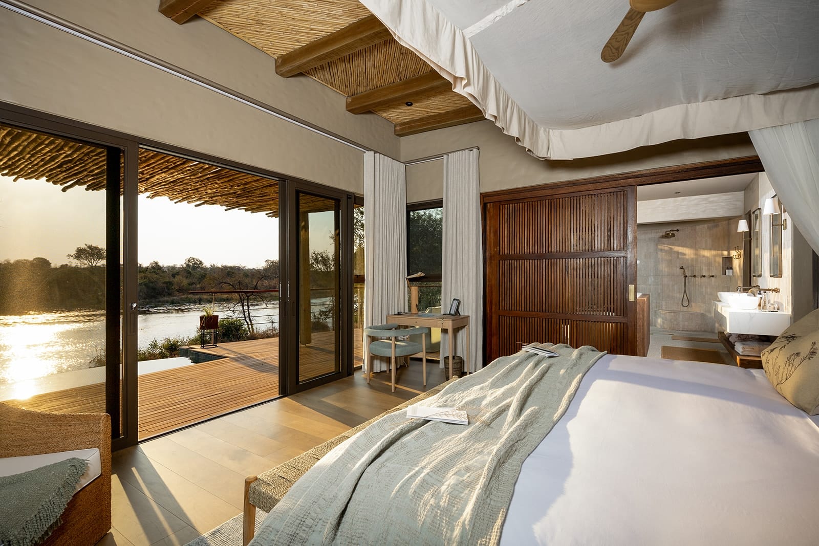 Inside a Shisaka Camp suite at Sandringham, a canopied bed and open glass doors framing the Timbavati River in low golden light