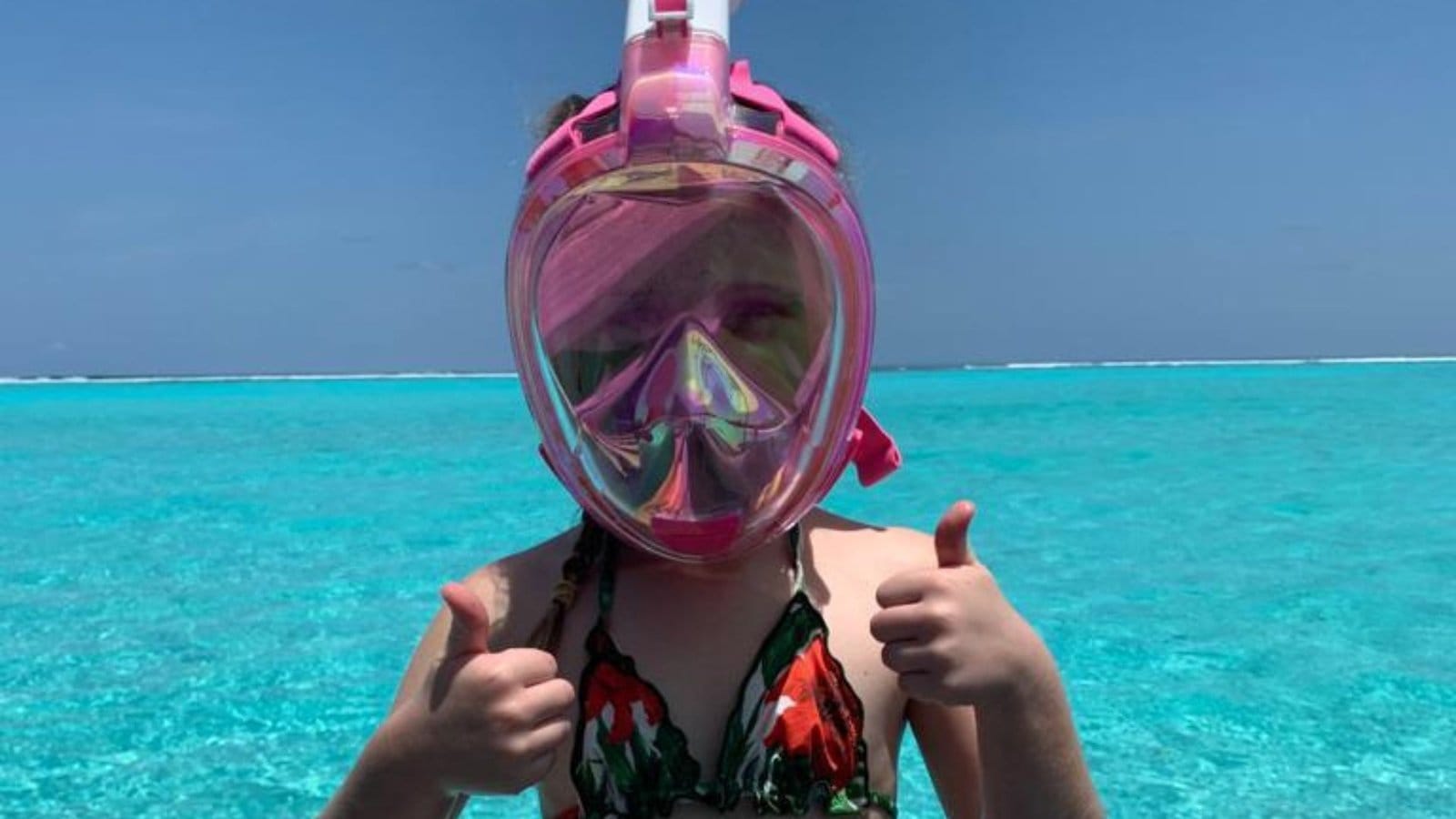 Young girl in a pink full-face snorkel mask giving two thumbs up above turquoise shallows