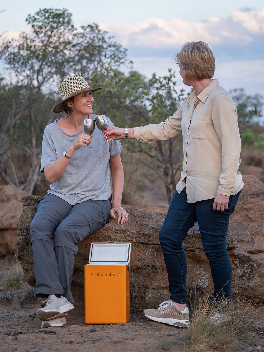Two women standing and sitting among rocks with drinks in hand at dusk in open bush