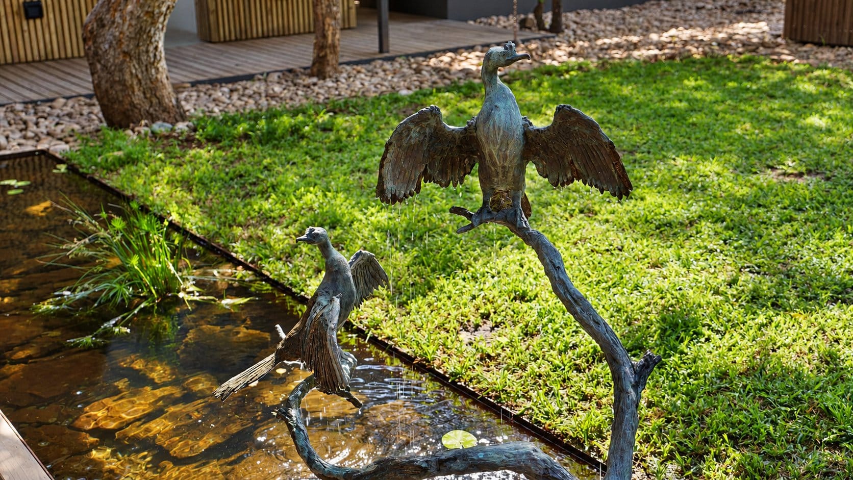 Two bronze cormorant sculptures on a branch above a narrow water channel beside a lawn