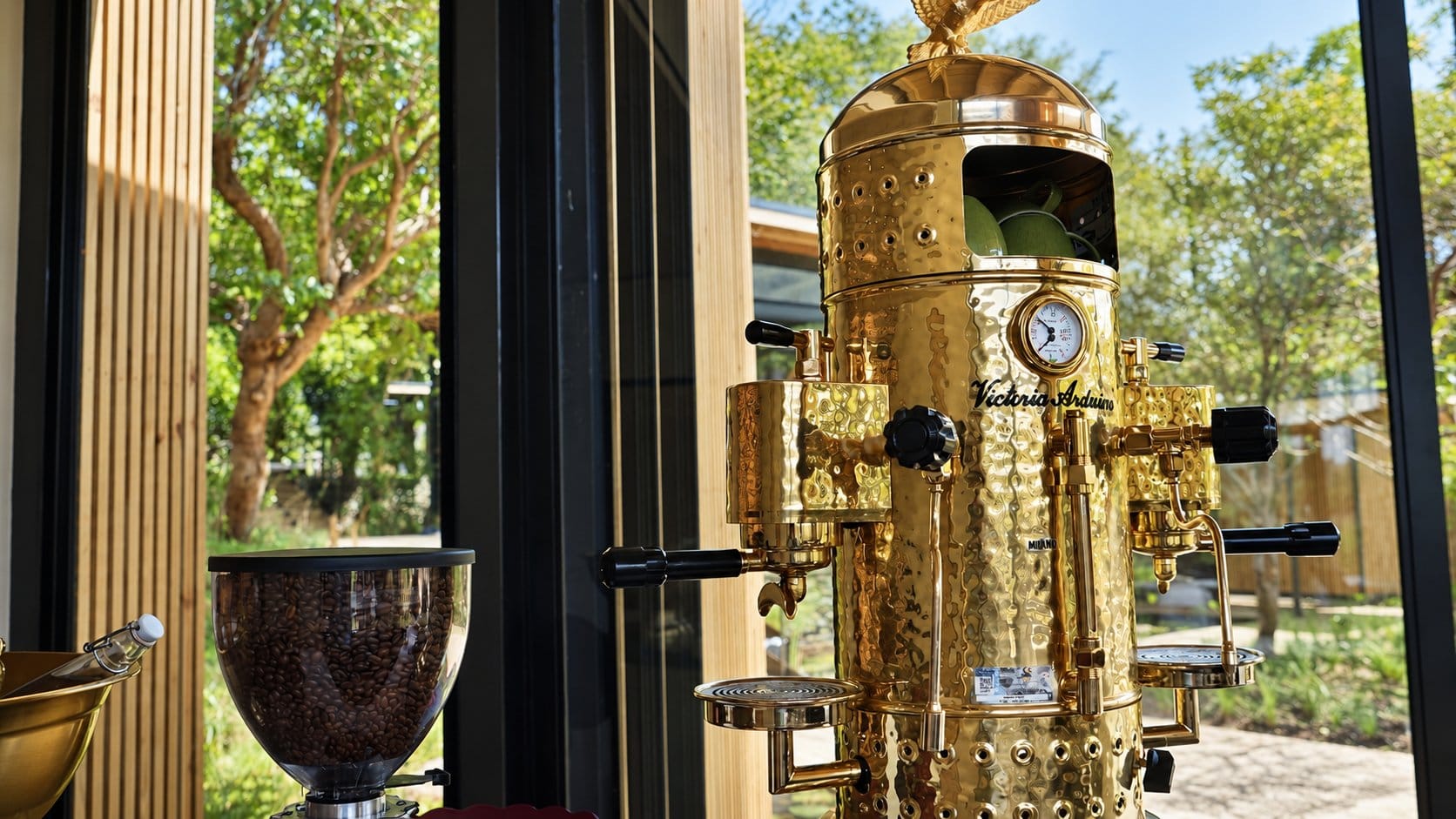 Hammered gold espresso machine topped with a gold eagle, beside a coffee grinder at a window onto the garden