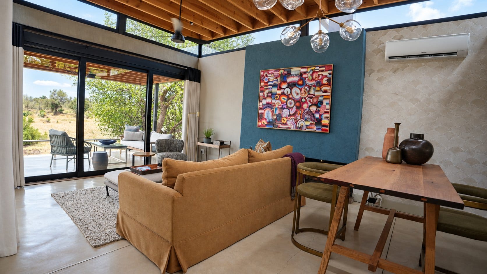 Suite lounge with a blue feature wall, bold abstract artwork, a mustard sofa and a timber dining table, with glass doors open to the bush