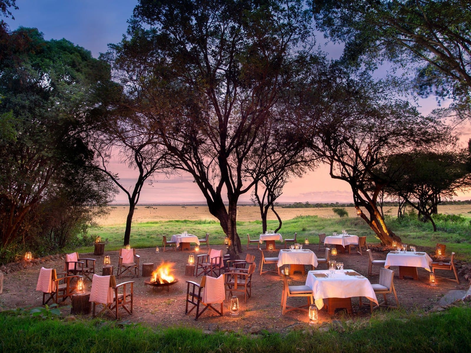 Firelit open-air dining under trees at a Masai Mara conservancy lodge at dusk, the plains beyond