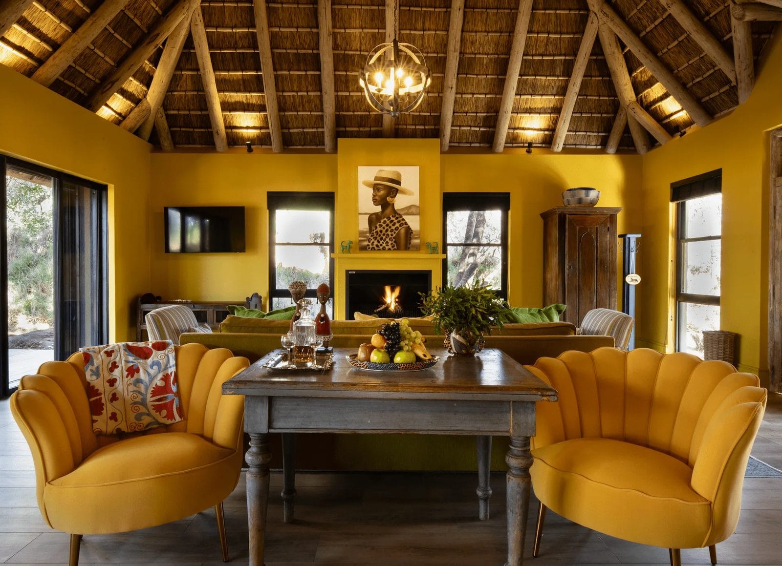Yellow lodge lounge under a thatched roof, with a fire lit, velvet armchairs and a table set with fruit