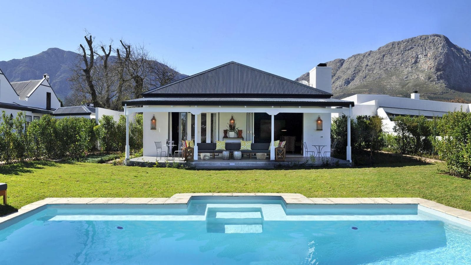 White cottage with a shaded veranda and a pool on the lawn beneath the Franschhoek mountains
