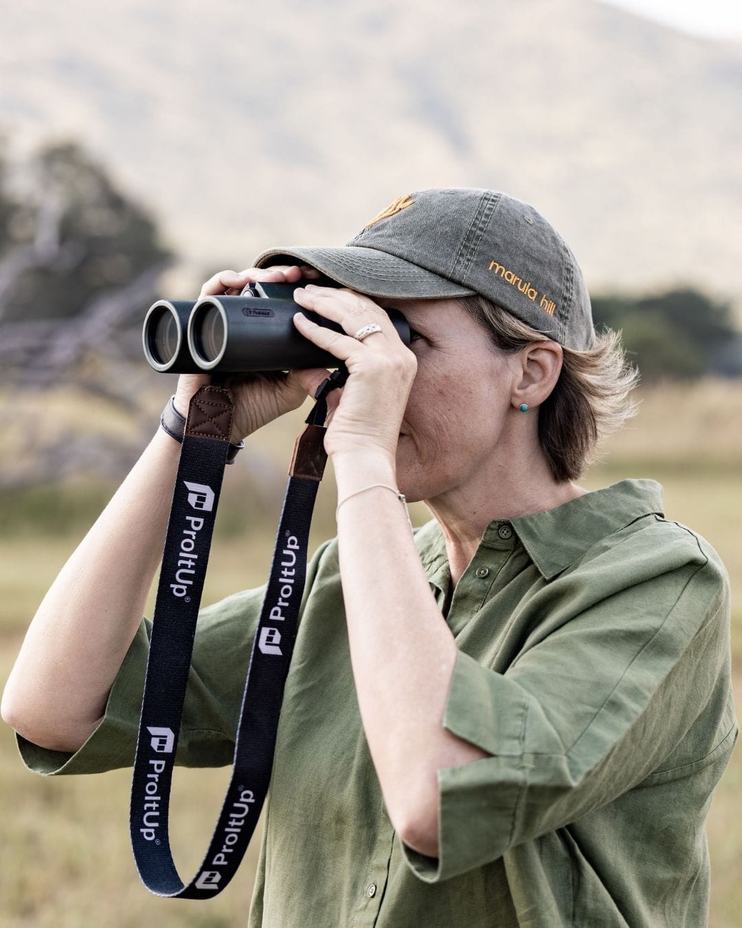 Sian Loehrer looking through binoculars in open grassland, wearing a Marula Hill cap