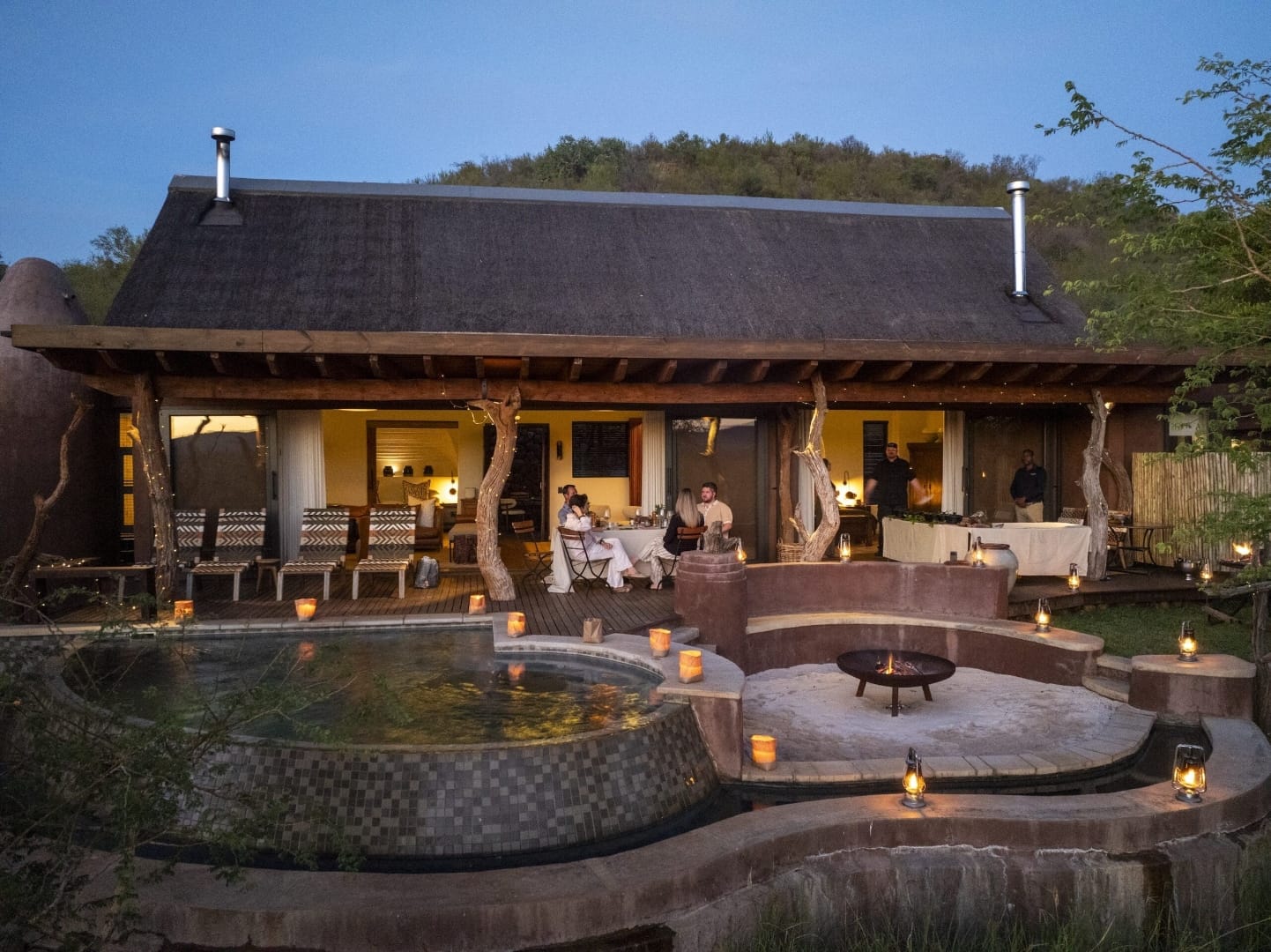Thatched private house lit by lanterns at dusk, with a pool, fire pit and a table set on the deck