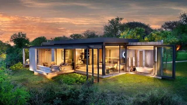 Glass-fronted safari villa lit from within at dusk, surrounded by bush