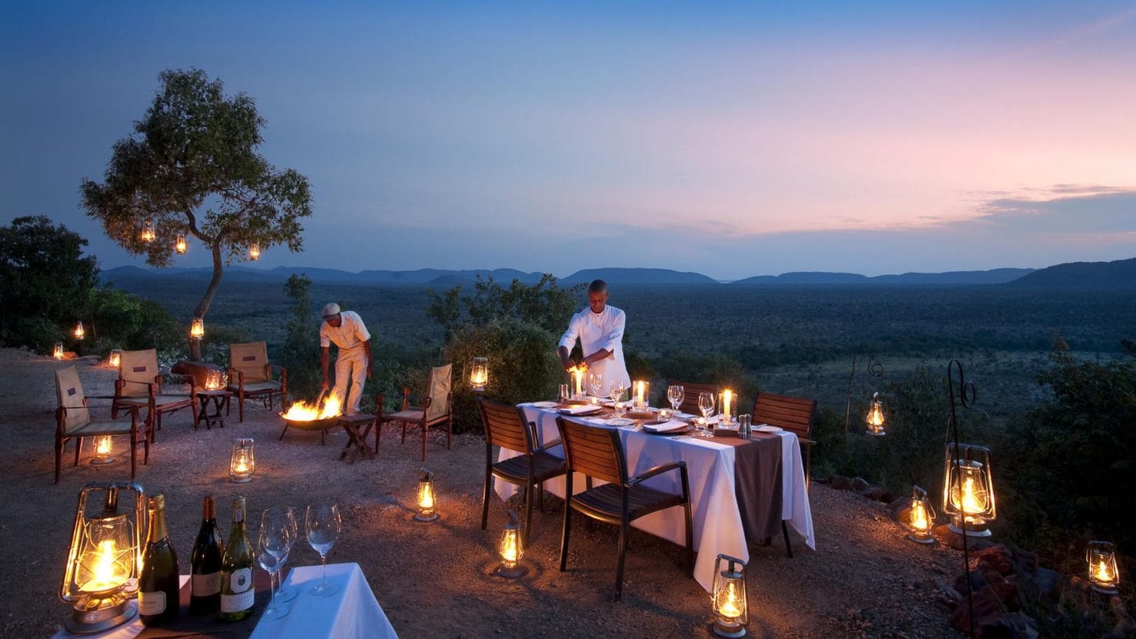 Lantern-lit dinner table set on a hilltop in Madikwe at dusk, with a fire burning nearby