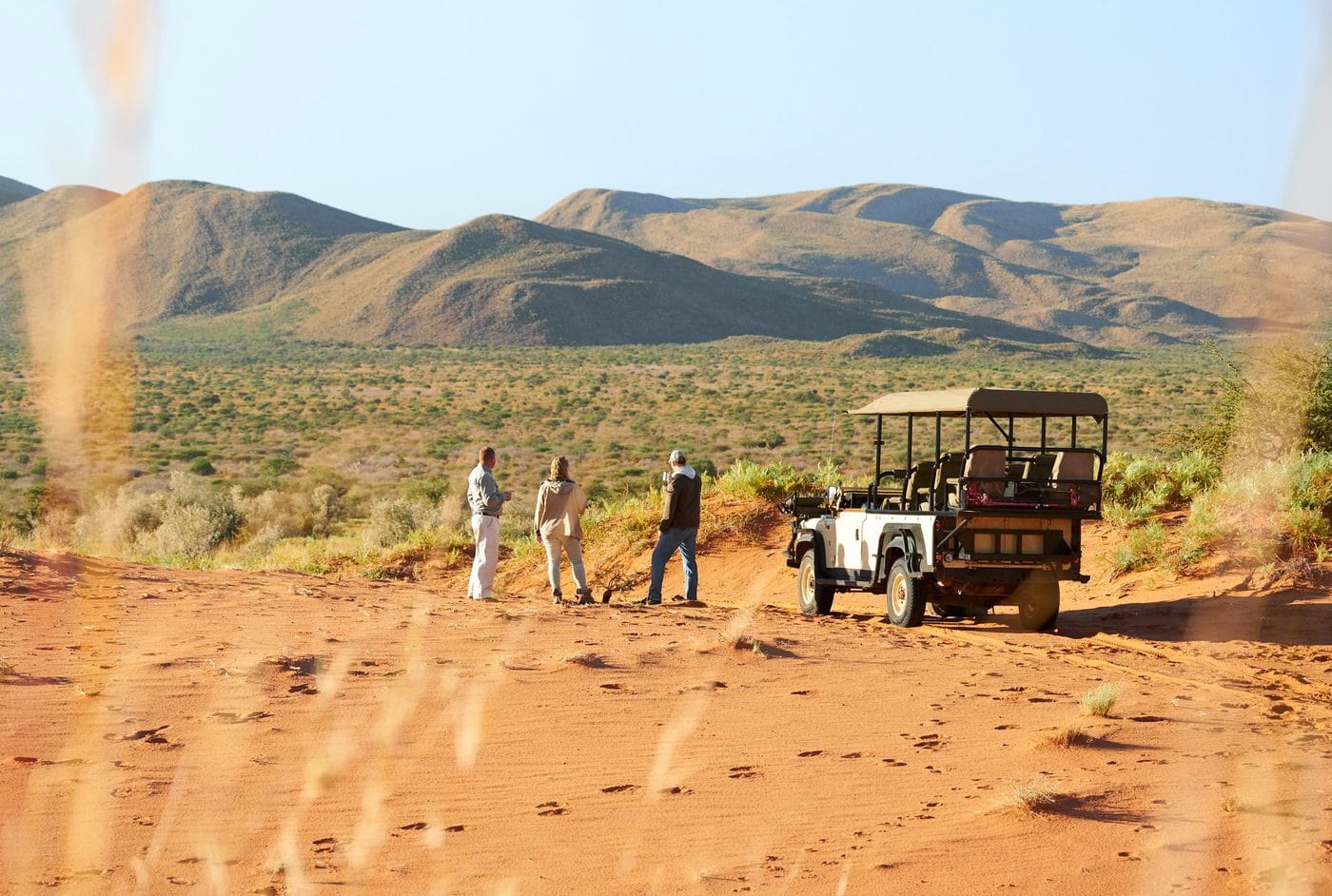 Guests standing beside an open game vehicle on red Kalahari sand with low hills behind