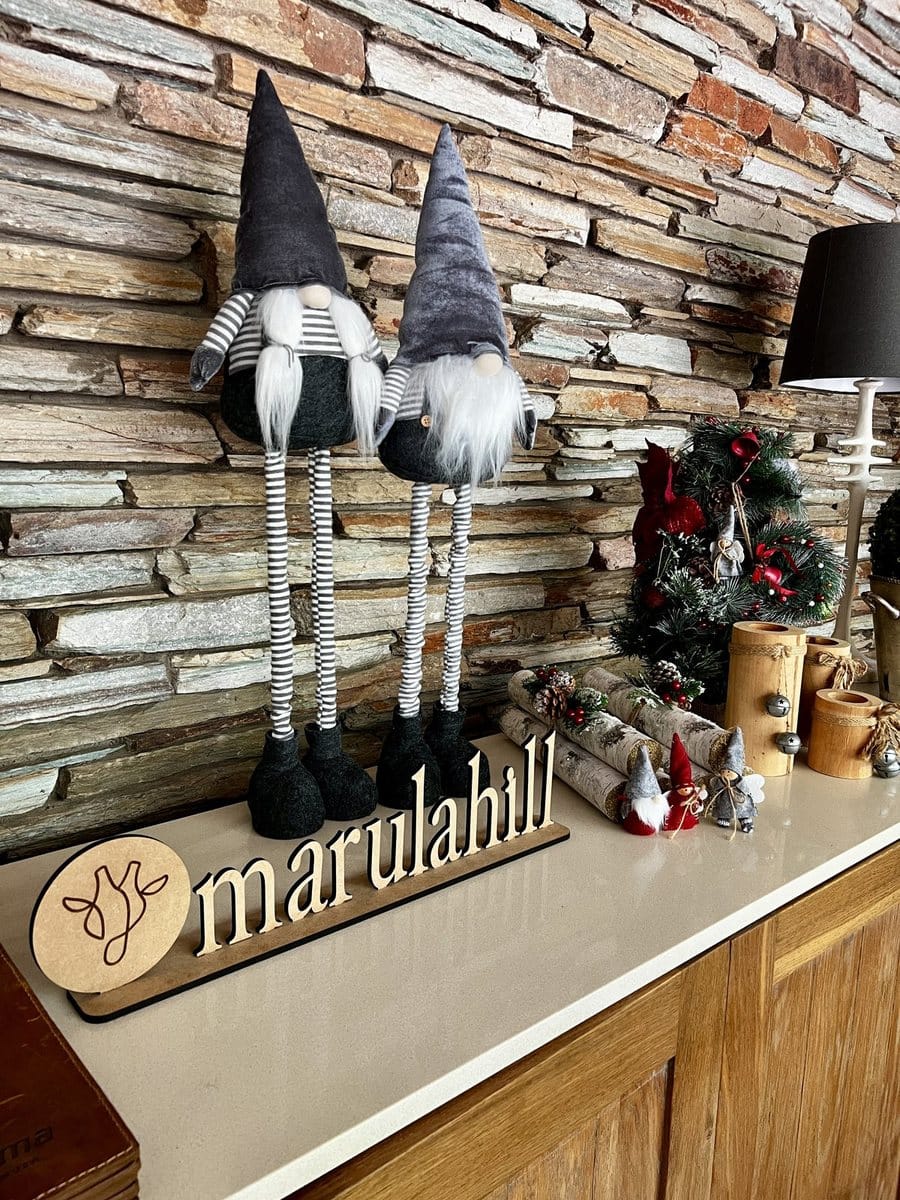 Christmas gnomes, a small decorated tree, birch logs and a wooden Marula Hill sign on a counter against a stone wall