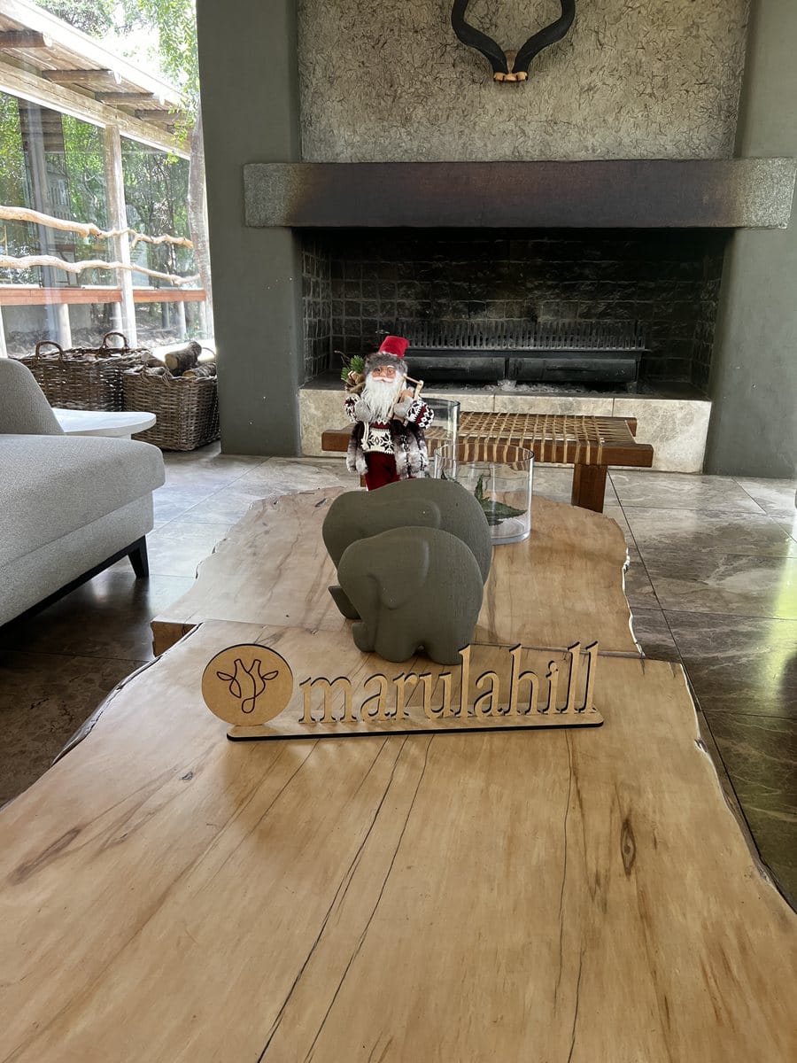 A small Father Christmas figure, two grey elephant ornaments and a wooden Marula Hill sign on a wooden table in a lodge lounge with a stone fireplace