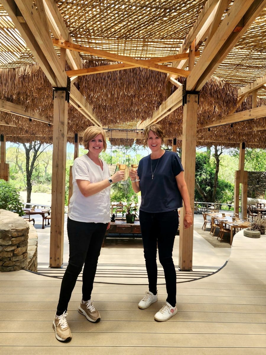 Vikki and Sian smiling and raising glasses of wine on a thatched lodge deck, with green summer bush behind them