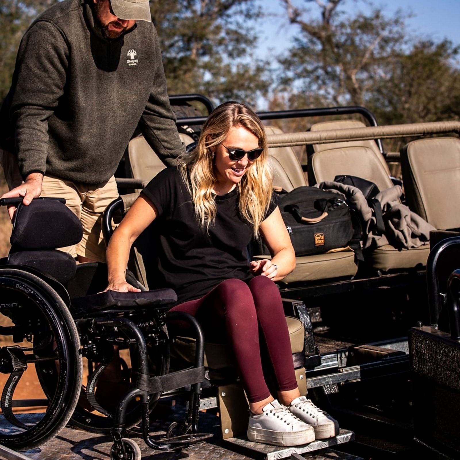 A woman transferring from her wheelchair onto an open safari vehicle, steadied by a guide in a Ximuwu Lodge fleece