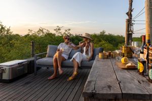 Two women on the deck of the Kingston Treehouse at Lion Sands, golden-hour light over the Sabi Sand bush, a sundowner set on the wooden table in front of them.