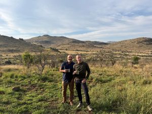 Two men holding enamel mugs on a grassy hillside in the bush, with low hills behind