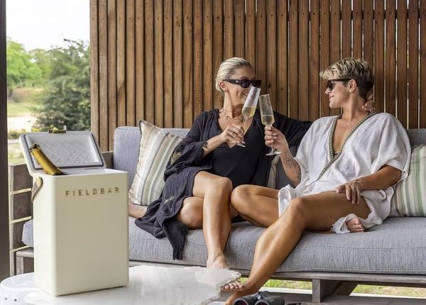 Two women raising glasses of sparkling wine on a shaded deck sofa overlooking the bush