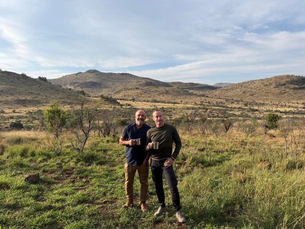 Two men holding enamel mugs on a grassy hillside in the bush, with low hills behind