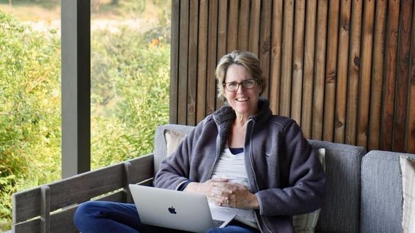 Sian Loehrer smiling with a laptop, sitting cross-legged on a covered outdoor daybed at Lion Sands Ivory Lodge, with green bush behind her