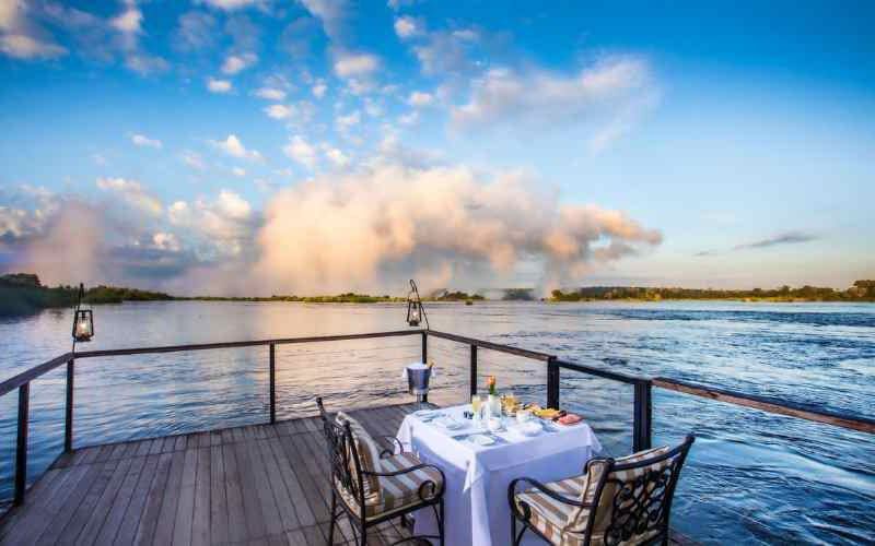 Private breakfast setup on a riverside deck overlooking the mist of Victoria Falls on the Zambezi River, with dramatic morning light and expansive sky.