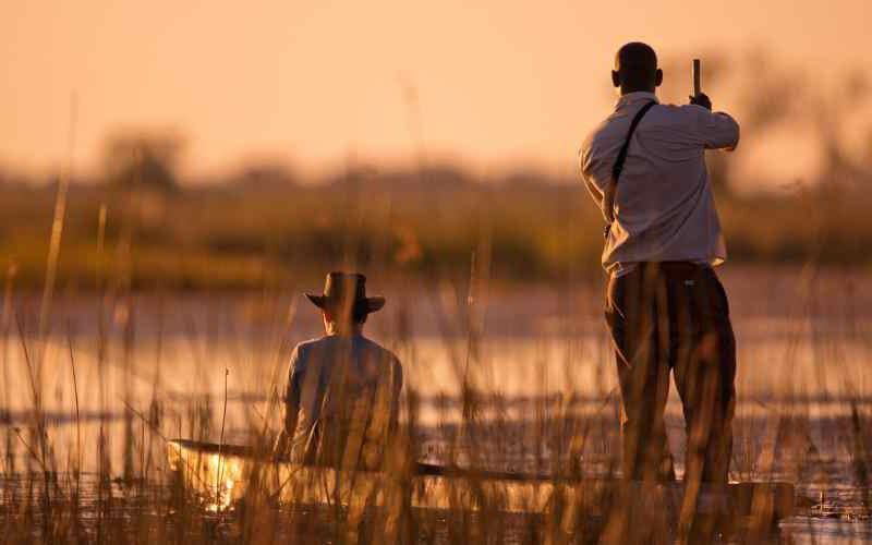 Solo traveller and local guide on a mokoro safari at sunset in the Okavango Delta, drifting through reeds during a luxury solo safari in Africa.
