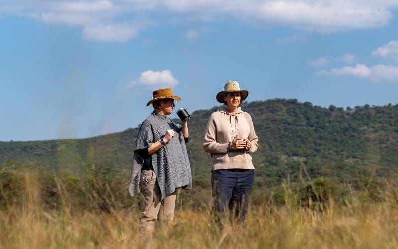 Sian Loehrer and Vikki Jackson enjoying coffee in the African bush, surrounded by golden grass and rolling hills—capturing the peaceful, personalised moments that define a Marula Hill Travel safari.