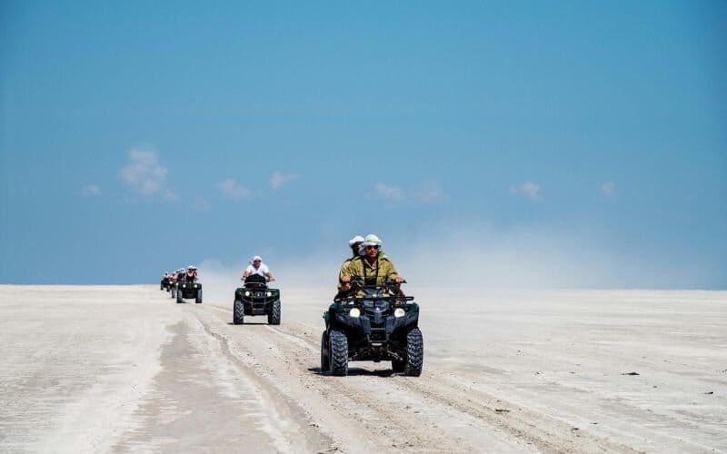 Group of travellers riding quad bikes across the white salt flats of Botswana’s Makgadikgadi Pans under a clear blue sky.