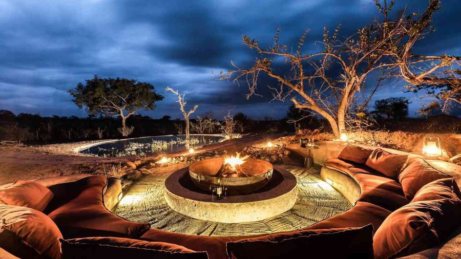 Luxury firepit lounge area at a Sabi Sand safari lodge, illuminated by lanterns under a dramatic night sky, overlooking a private plunge pool and the African bush.