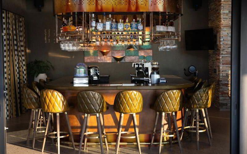  The elegant indoor bar at Ximuwu Lodge, featuring a U-shaped wooden counter surrounded by luxurious quilted leather bar stools in mustard and olive tones. Overhead, a suspended rack displays premium spirits and glassware. The space is styled with modern lighting, textured wall art, and natural stone and fabric accents, capturing Ximuwu’s signature blend of contemporary design and safari chic.